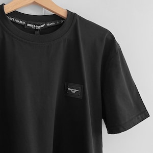 May include: A black crew neck t-shirt with a small rectangular label on the chest. The label has text on it. The neckline has the text "DOLCE & GABBANA" printed on it. The shirt is hanging on a wooden hanger.