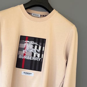 Può includere: Felpa beige con una grande toppa quadrata con il logo Burberry e un cavaliere a cavallo. La toppa presenta un motivo a quadri neri, rossi e bianchi. Il logo Burberry è stampato anche sul girocollo.