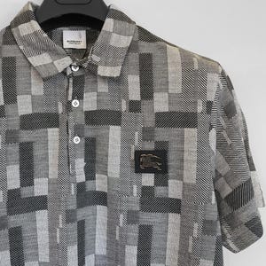 May include: A short-sleeved polo shirt with a geometric pattern in shades of gray and white. The shirt features a collared neckline, button closure, and a small logo patch on the chest. The Burberry logo is visible on the label.