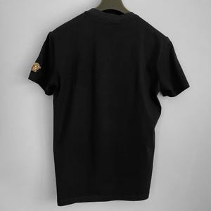 May include: Black short-sleeved t-shirt with a gold-colored emblem on the left sleeve. The shirt is hanging on a black hanger against a plain white background. The emblem appears to be a Medusa head.