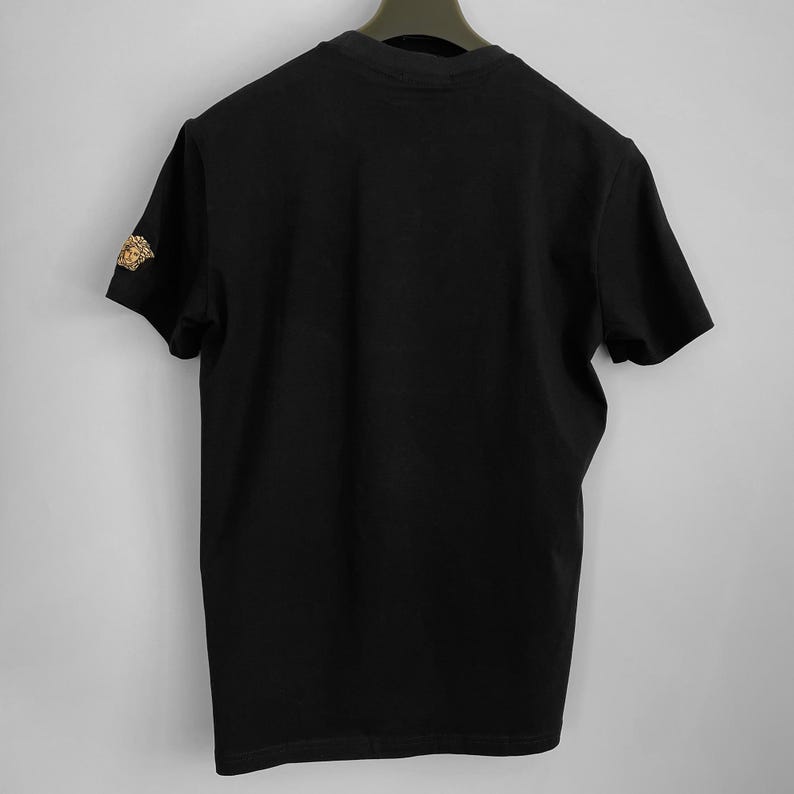 May include: Black short-sleeved t-shirt with a gold-colored emblem on the left sleeve. The shirt is hanging on a black hanger against a plain white background. The emblem appears to be a Medusa head.