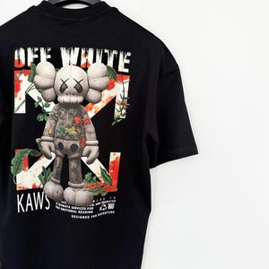 Puede incluir: Camiseta negra con un diseño de figura KAWS con detalles florales y los textos "OFF WHITE" y "KAWS". El diseño incluye flechas blancas y el texto "DESIGNED FOR ADVENTURE".