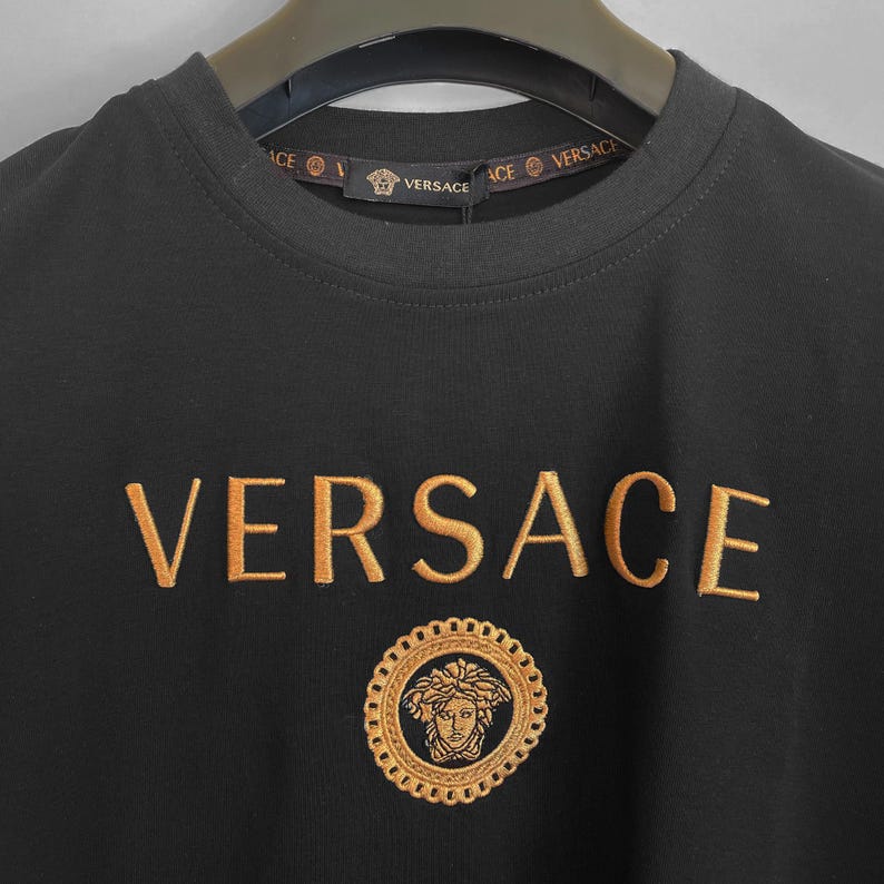 May include: Black crew neck t-shirt with gold embroidered "VERSACE" across the chest. Below the text is a gold embroidered circular emblem with a face in the center. The neckline has the brand name printed in gold.