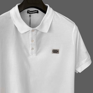 May include: White polo shirt with a classic collar and short sleeves. Features a three-button placket and a small rectangular logo on the chest. The shirt is made of a smooth, likely breathable fabric, and is hanging on a black hanger.