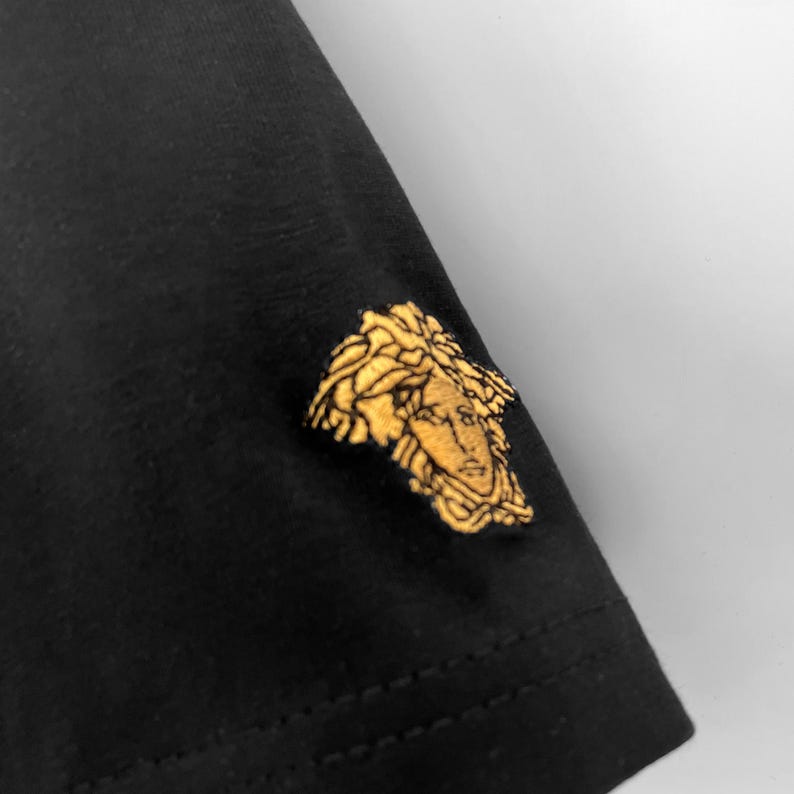 May include: Black t-shirt with a gold-colored Medusa head emblem on the sleeve. The emblem is intricately detailed, featuring a profile of a woman's face with flowing hair. The shirt's fabric appears smooth and the emblem is stitched on.