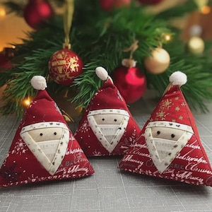May include: Three handmade Christmas ornaments shaped like Santa Claus hats. The red fabric hats have white trim, a white pom-pom, and a stitched face. The background includes a Christmas tree and red and gold ornaments.