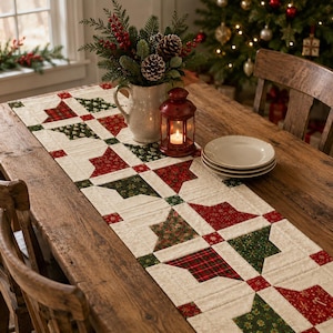 May include: A rectangular quilted table runner featuring a repeating pattern of red and green fabric blocks with snowflake and plaid prints on a cream background. The runner is laid flat on a wooden table with a white pitcher and a red lantern.