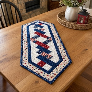 May include: A patriotic table runner with a quilted design. The runner features a white center with red and blue zigzag stripes, incorporating star and stripe patterns. A blue border with star accents completes the design. This decorative piece is perfect for adding a festive touch to any table.