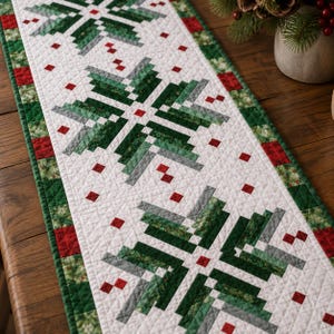 May include: A rectangular quilted table runner featuring a white background with a repeating geometric star pattern in shades of dark green, light green, and grey. The runner is bordered by a patchwork of green and red squares and small red accents.