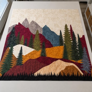 May include: A rectangular quilt featuring a landscape scene with layered mountains in shades of gray, blue, and deep red. The foreground includes evergreen trees in various shades of green and black, set against rolling hills of gold and tan fabric.