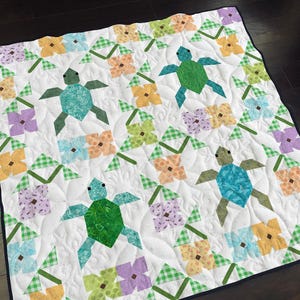 May include: A white quilt with a green, blue, and yellow floral pattern. The quilt features a repeating design of stylized turtles with a green, blue, or yellow shell.