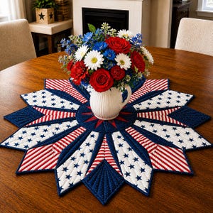 May include: A round wooden table with a patriotic starburst table runner. The runner features a blue, red, and white color scheme with stars and stripes. A white vase holds a bouquet of red roses, white daisies, and blue flowers.