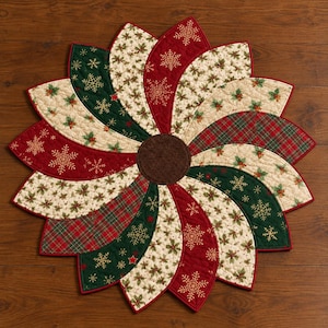 May include: A circular quilted table mat featuring a pinwheel design with alternating fabric segments in red, dark green, and cream. The patterns include gold embroidered snowflakes, holly leaves, and a red and green plaid, centered with a brown circle.