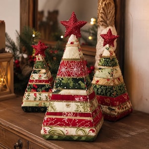 May include: Three cone-shaped fabric Christmas tree decorations featuring horizontal patchwork strips in red, green, and cream patterns. Each tree is topped with a small, glittery red star. The fabric pieces are joined with gold-toned stitching.