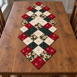 May include: A diamond-shaped quilted table runner featuring a patchwork design of cream squares with gold snowflake patterns, dark green plaid squares, and red squares with white snowflake and holly berry prints arranged in a repeating pattern.