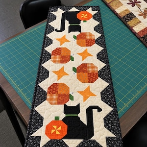 May include: A rectangular quilted table runner featuring a cream-colored center panel with alternating black cat silhouettes, orange patchwork pumpkins, and orange four-pointed stars, all bordered by a black floral fabric with white sawtooth edges.