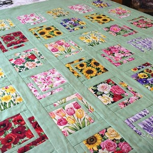 May include: A floral quilt featuring a patchwork design with various flower patterns, including sunflowers, roses, and tulips. The quilt has a light green background and is made of fabric squares. The overall design is a mix of colors, including red, yellow, pink, and purple.