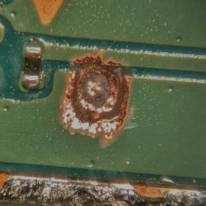 May include: A close-up view of a green printed circuit board featuring copper-colored conductive traces, a small orange triangle symbol, and a circular area showing signs of oxidation or corrosion with white residue near a metallic component.