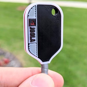 May include: A black and white key tag with the word "Joola" printed on it. The tag has a rounded rectangular shape with a hole at the top. The tag is held by a gray wire.