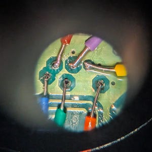 Könnte beinhalten: Eine kreisförmige Ansicht einer grünen Leiterplatte mit sechs gelöteten Drahtverbindungen. Die Drähte sind in Rot, Violett, Gelb, Orange, Grün und Blau isoliert und jeweils an einem metallischen Kontaktpunkt auf der Platinenoberfläche befestigt.