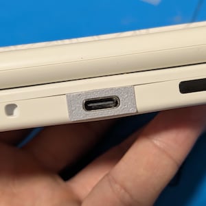 May include: A white electronic device featuring a USB-C charging port integrated into the side casing. The port is surrounded by a textured, light gray rectangular frame that sits flush against the smooth, off-white plastic surface of the device.