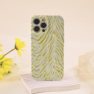 Puede incluir: Una funda para smartphone con un patrón repetitivo de rayas de cebra en verde oliva y gris claro. La funda tiene un recorte para un sistema de cámara de triple lente en la esquina superior izquierda, mostrando tres lentes circulares.