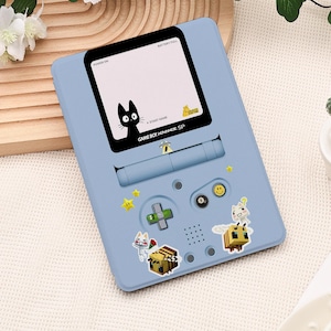 May include: A pale blue Game Boy Advance SP handheld console with a black screen showing a black cat illustration. The console is adorned with stickers of stars, a cross, and pixelated bees and other characters.