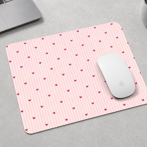 Peut inclure: Un tapis de souris rectangulaire présentant un motif à rayures verticales rose clair et blanc, orné de petits cœurs rouges dispersés. Une souris d'ordinateur blanche repose sur le côté droit de la surface du tapis.
