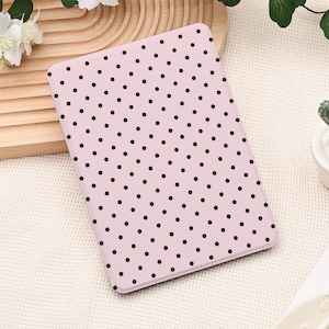 Puede incluir: Una funda para tableta de color rosa claro con un patrón repetido de pequeños lunares marrones oscuros. La funda rectangular está hecha de un material liso. El diseño es sencillo y femenino.
