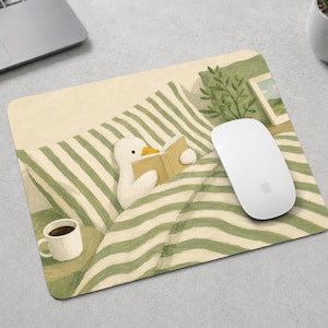 Puede incluir: Una alfombrilla de ratón rectangular con la ilustración de un pato blanco de pico naranja leyendo un libro en una cama con sábanas de rayas verdes y blancas. Una taza de café blanca está cerca y un ratón de ordenador blanco descansa encima.