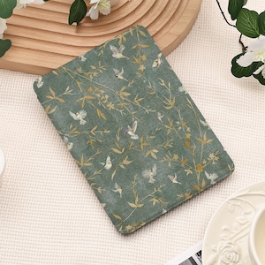 May include: A rectangular e-reader cover featuring a muted sage green background with a repeating pattern of small white birds and delicate golden-yellow branches and leaves. The cover has a flat, smooth surface and rounded corners.