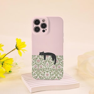 May include: A light pink phone case featuring a black cat design. The cat is lying down above a floral pattern with pink flowers and green leaves. The case is resting on a white, rectangular stand.