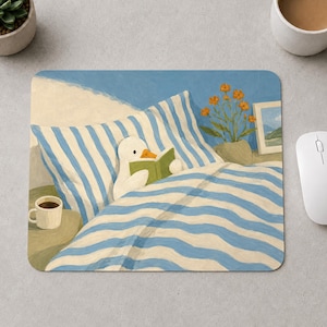 Peut inclure: Un tapis de souris rectangulaire illustrant un canard blanc au bec orange lisant un livre vert dans un lit aux draps rayés bleus et blancs. Une petite tasse blanche est posée sur une table de chevet à côté du canard.