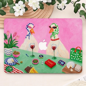 May include: A rectangular laptop skin featuring a vibrant pink background with two white geese wearing headscarves and sunglasses. They sit at a green table with wine glasses, a red box labelled SASSY, a green bag, cheese, and chocolate.