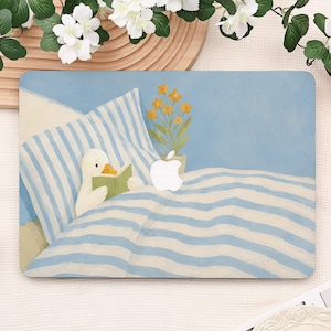 Puede incluir: Una pegatina para portátil con una ilustración pintada de un pato blanco leyendo un libro verde en una cama de rayas azules y blancas. Una pequeña planta en maceta con flores amarillas se sitúa junto al logotipo blanco de Apple.
