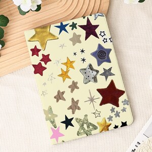 May include: A cream-colored notebook cover adorned with various star designs in gold, red, blue, and silver. The stars vary in size and texture, creating a whimsical and artistic pattern. The cover is rectangular and appears to be made of a smooth material.