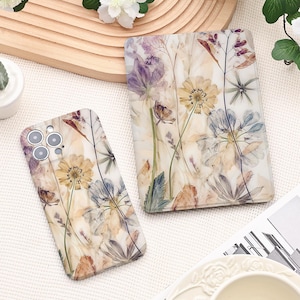 May include: A phone case and a rectangular card holder featuring a floral design with pressed flowers in shades of yellow, purple, and white. The case has camera cutouts. The card holder and case are set against a white background.