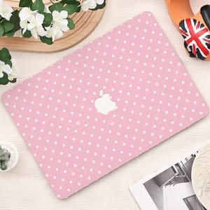 May include: A light pink laptop cover featuring a repeating pattern of small white polka dots. The cover has a central cutout revealing the silver Apple logo. The design is flat and rectangular, covering the top surface of the portable computer.