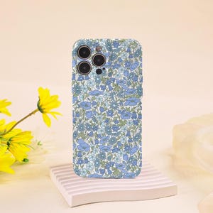 Op de afbeelding: Een telefoonhoesje met een bloemenpatroon in blauw en wit. Het patroon toont blauwe en lichtblauwe bloemen op een witte achtergrond. De hoes wordt gepresenteerd op een witte, geometrische standaard.
