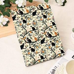 May include: A rectangular notebook with a cream-colored background featuring a repeating pattern of black, orange, and white cats perched on tree branches with orange fruit and green leaves. The notebook is open.