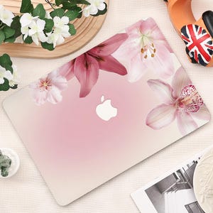 以下が含まれることがあります： ピンク色のノートパソコン。ピンクと白のユリと蘭の花柄のデザイン。Appleのロゴが見えます。花はノートパソコンの上部に配置されています。