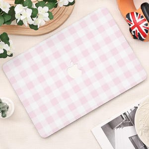 Könnte beinhalten: Eine Laptop-Hülle mit rosa und weißem Vichy-Muster und dem Apple-Logo-Ausschnitt. Die Hülle liegt auf einer hellen Oberfläche, mit dekorativen Elementen wie Blumen und einem kleinen Spielzeugauto.