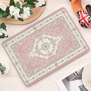 Puede incluir: Alfombra rectangular rosa con un diseño floral y de volutas detallado en crema y gris claro. La alfombra tiene un borde con detalles florales y un patrón de medallón central. La alfombra es probablemente para uso decorativo o funcional.