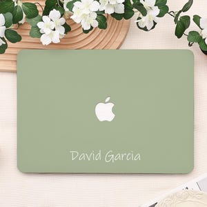 May include: A sage green laptop case with the Apple logo and the name "David Garcia" in white script. The case is on a light-colored surface, with white flowers and green leaves in the background.