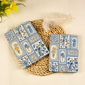 May include: Two blue and white patterned journals with a tropical design featuring birds and plants. The journals are on a woven straw surface. The text "Hudson" is printed on one of the journals.