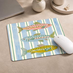 May include: A rectangular mouse pad featuring a watercolour-style pattern of three fish in pink, blue, and yellow, each decorated with small green spots. The background consists of vertical stripes in alternating shades of light blue and cream.