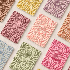 Op de afbeelding: Een collectie rechthoekige paspoorthouders met een zebrastreepmotief in diverse kleuren zoals geel, paars, roze, oranje, blauw en groen. Elke houder is gepersonaliseerd met tweeletterige initialen zoals WE, HY, EM, SE, EG en NM.