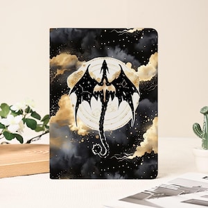 May include: A black and gold journal featuring a dragon design. The dragon is silhouetted against a white circle with a starry sky and gold clouds in the background. The journal has a matte finish.