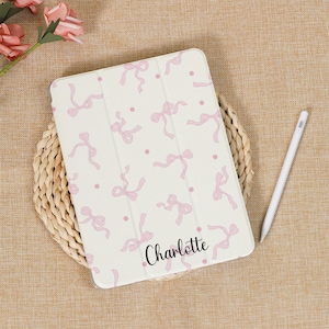 以下が含まれることがあります： ピンクのリボン柄が施された白いiPadケース。前面には「Charlotte」という名前が印刷されています。