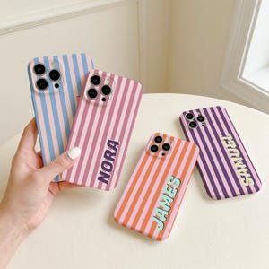 May include: Four striped phone cases in various colours, including blue, pink, orange, and purple. The cases feature the names "NORA", "JAMES", and "SAMUEL". One case is held by a hand. The cases have a vertical stripe design.